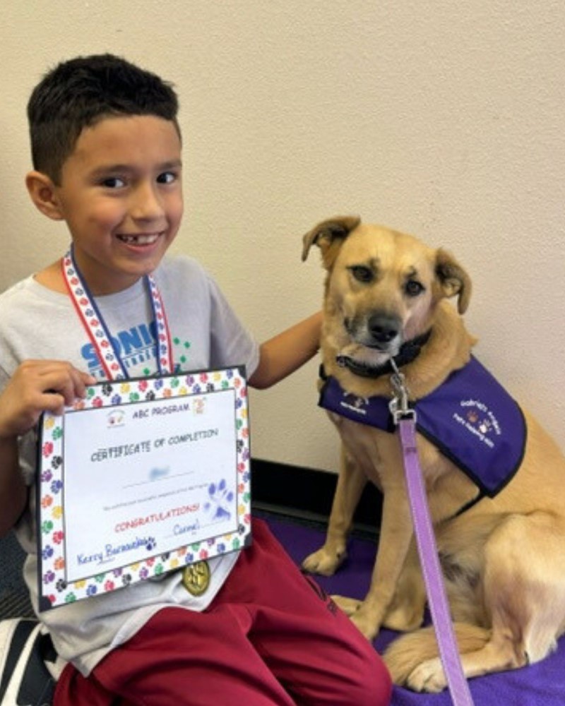 A little boy holds his certificate of completion for the ABC Pet Therapy Program and poses next to Therapy Dog Carmel.