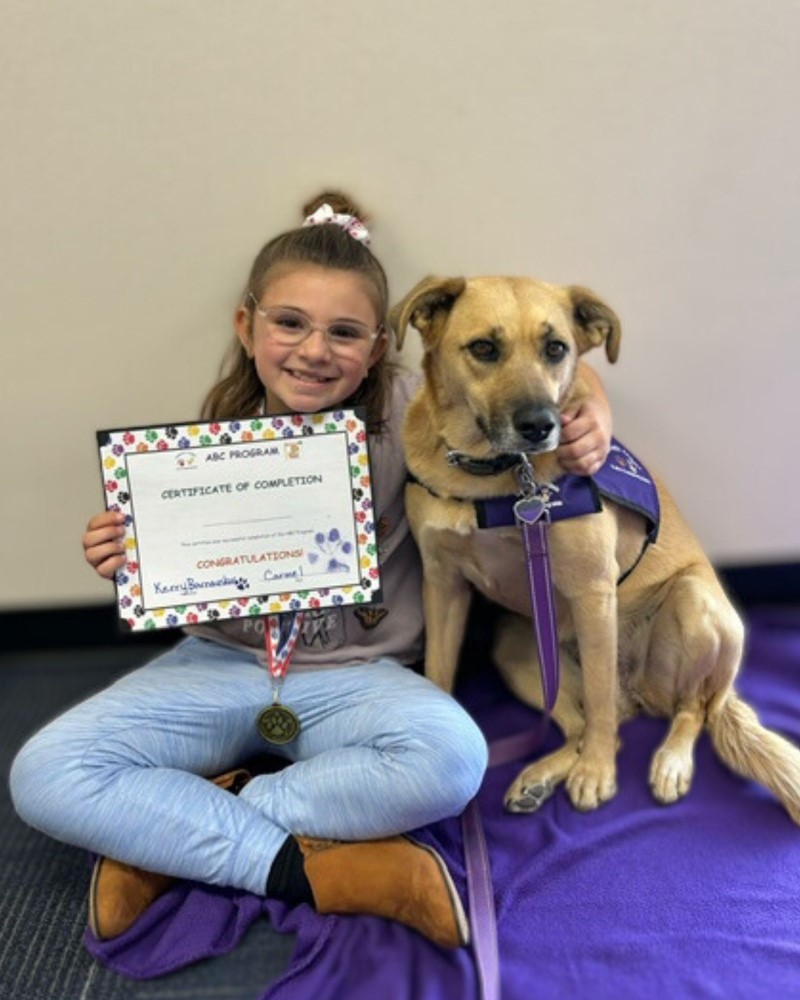 A little girl poses with Carmel while holding her certificate of completion from the ABC Pet Therapy Program.